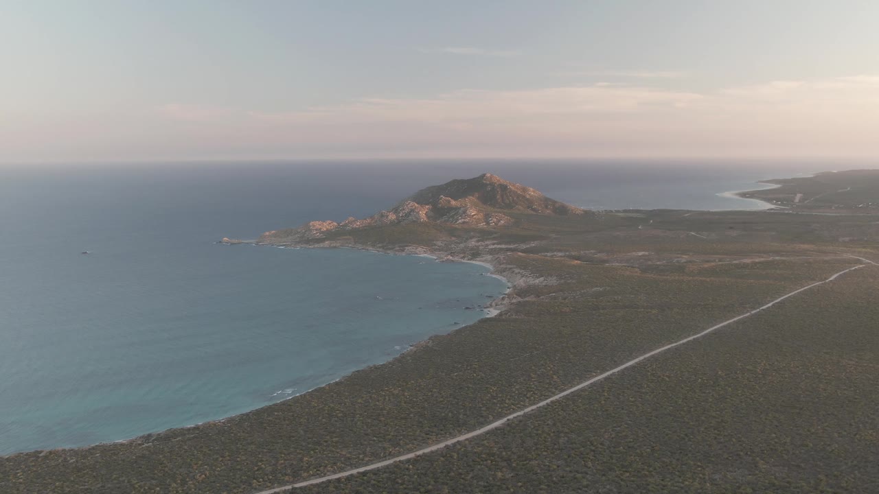 Wide Cabo Pulmo aerial frame with calm ocean at left, shoreline detail near the hill, the pale road stretching through scrubland, a rocky ridge beyond, and no visible people.