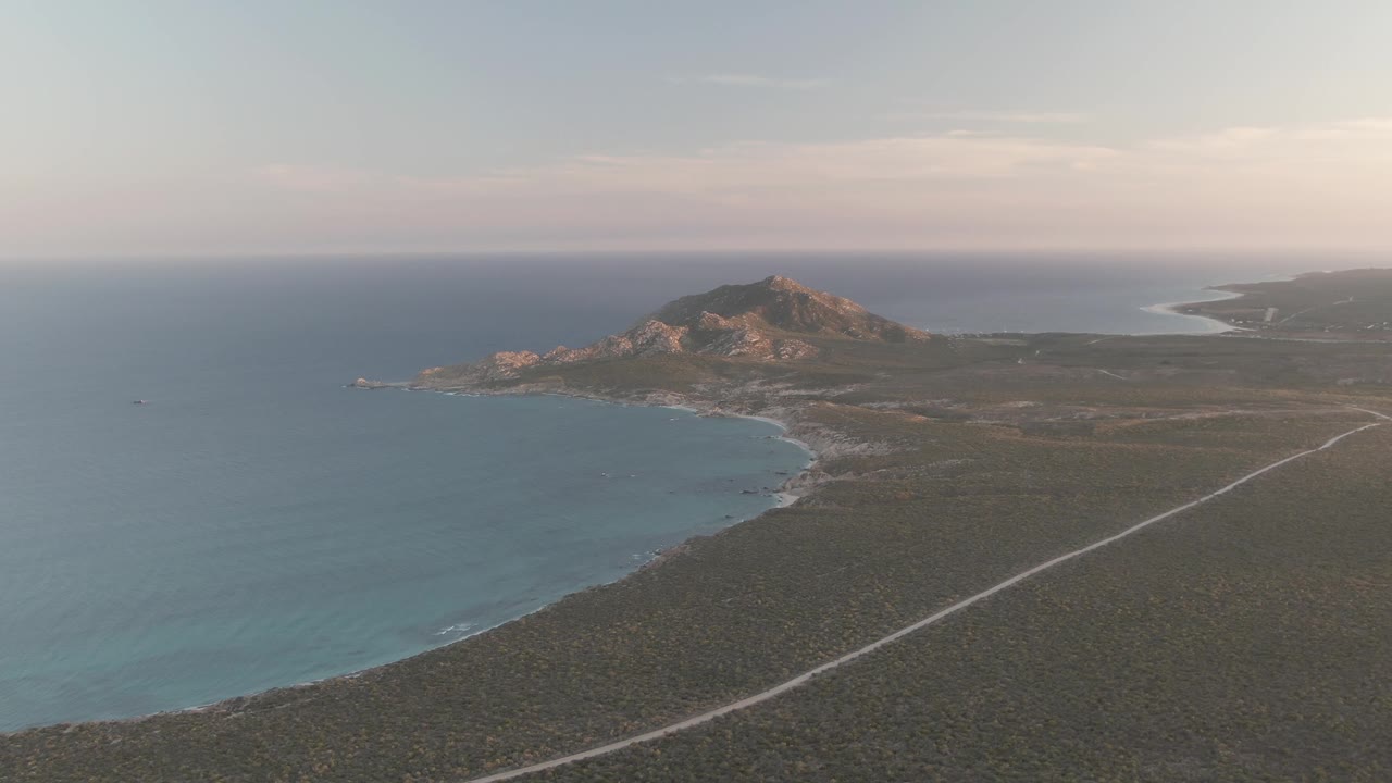 Aerial Cabo Pulmo coastline at the midpoint of the clip, showing blue water, open scrub terrain, a pale road angling toward distant rocky hills, layered horizon clouds, and no visible people.