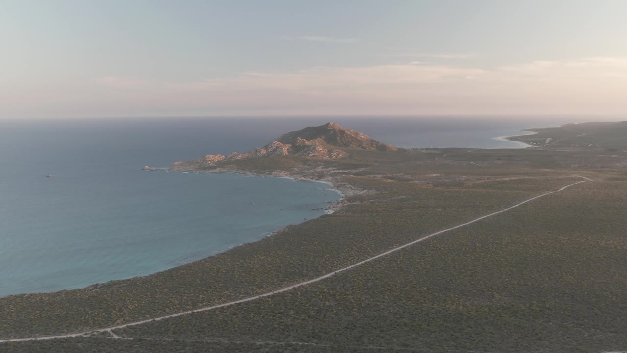 A high aerial view in Cabo Pulmo shows the coastline sweeping toward a rocky hill, with the road crossing low vegetation and no visible people.