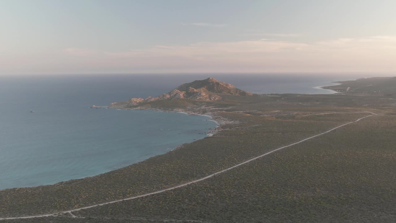 The Cabo Pulmo coastline remains wide in frame, with blue water at left, a pale road cutting through scrubland, a rocky hill near the center, and no visible people.