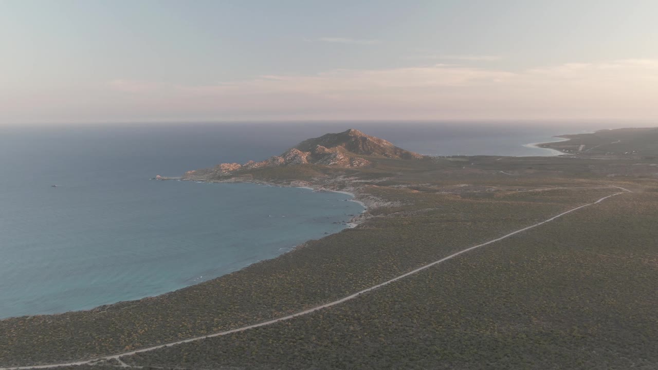 Mid-clip aerial frame from Cabo Pulmo with the road more prominent across the lower foreground, ocean and beach to the left, rocky hill beyond, and no visible people.