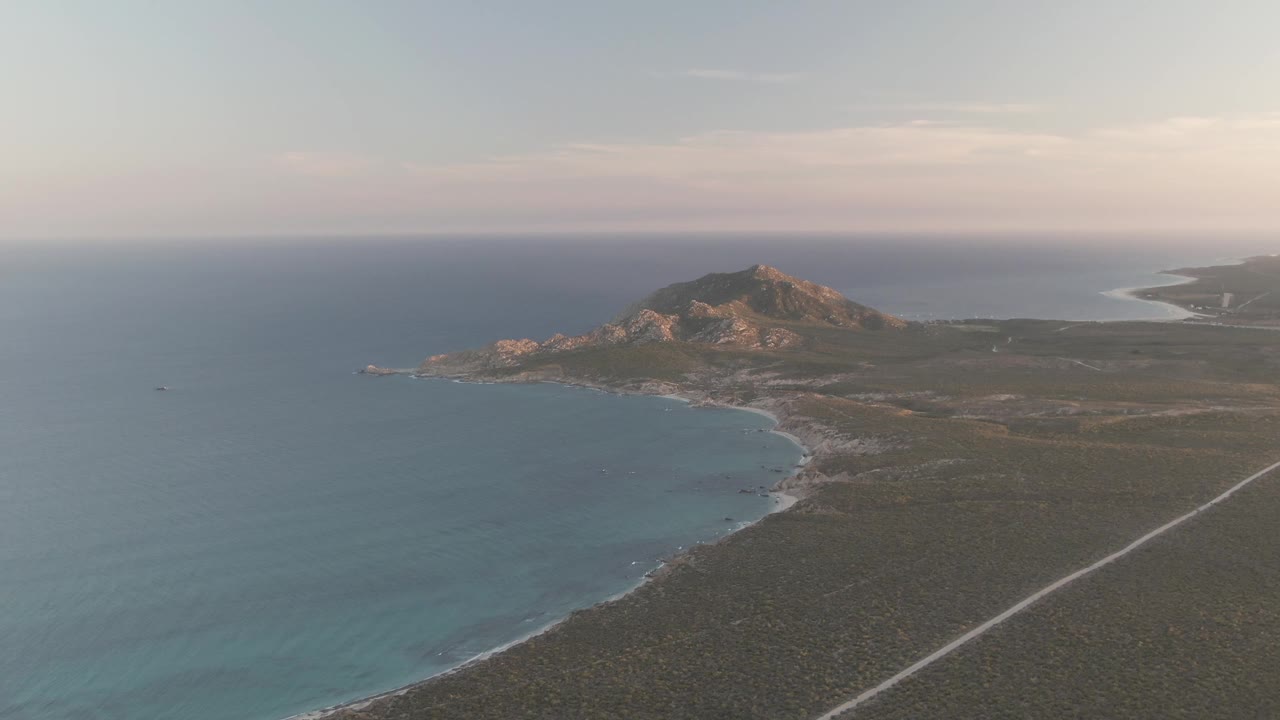 Opening aerial frame over Cabo Pulmo, Mexico, with blue ocean on the left, a curved shoreline below, dry coastal land, a pale road, distant hills, and no visible people.