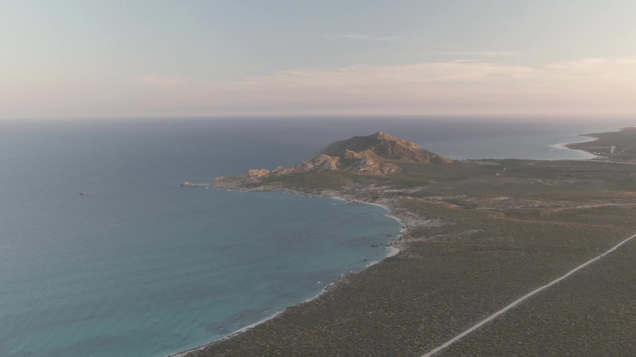 Second aerial frame over Cabo Pulmo, Baja California Sur, showing turquoise shallows along the coast, darker open water, scrubland, a straight road, distant hills, and no visible people.