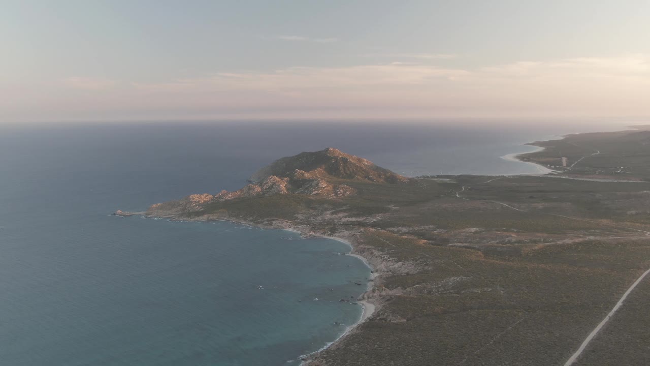 Aerial still of Cabo Pulmo showing open ocean, a rugged hill lit by low sun, rocky shoreline below, a pale road inland, and no visible people.