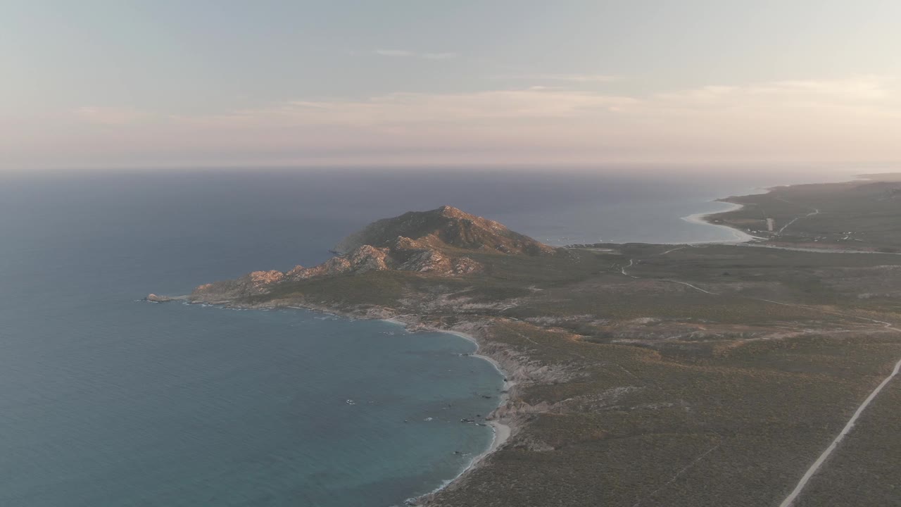 Cabo Pulmo coastline from above with turquoise shallows near the rocky shore, the hill set back from the water, a straight road at right, and no visible people.