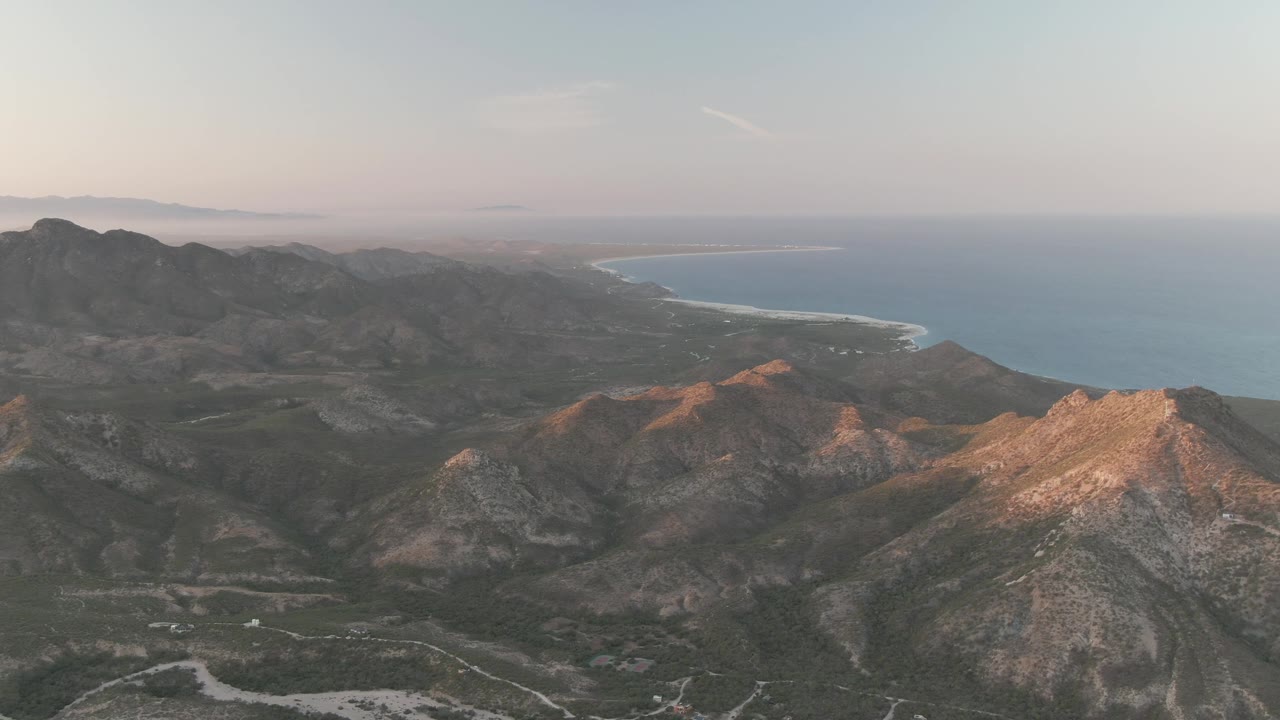 Early frame of Cabo Pulmo, Baja California Sur showing rugged mountain ridges in warm light with a sandy coast and blue ocean beyond, no visible people.