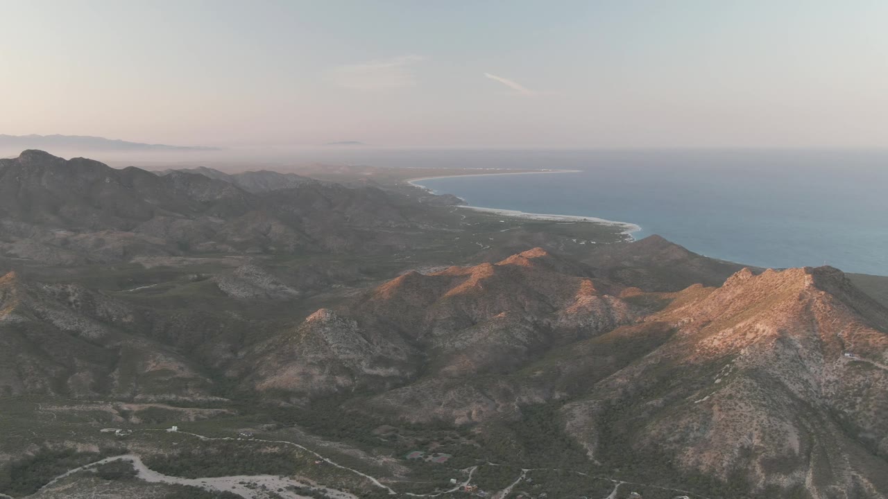 Cabo Pulmo frame with layered mountain slopes across the foreground, warm highlights on ridge tops, and a broad blue ocean horizon in the background, no visible people.