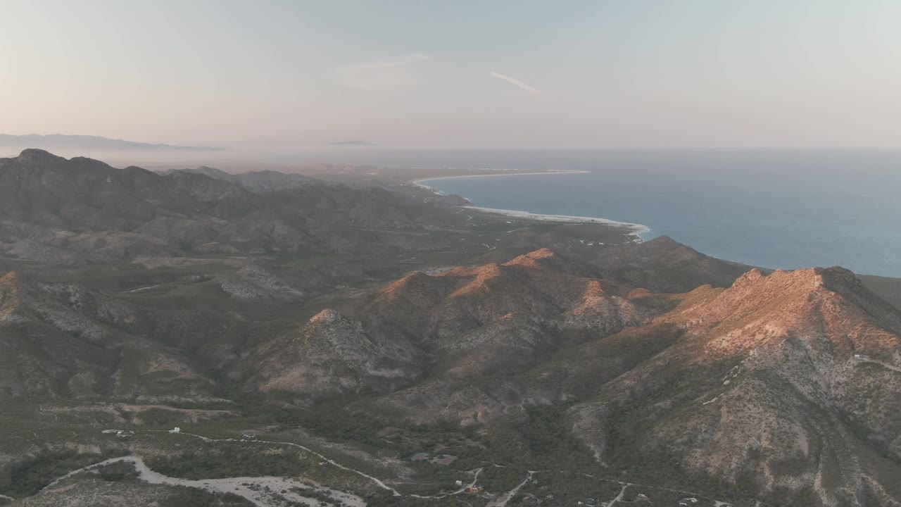 Mid-clip Cabo Pulmo view where the rocky foreground drops into scrubby valleys and a pale sandy shoreline traces the edge of the blue ocean, no visible people.