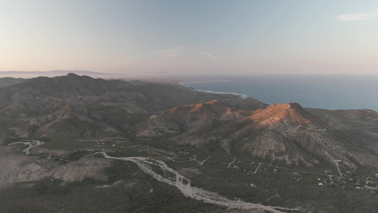 Cabo Pulmo aerial frame showing the sunlit central ridge brighter than the surrounding slopes, with dry valley roads below, ocean in the distance, and no visible people.