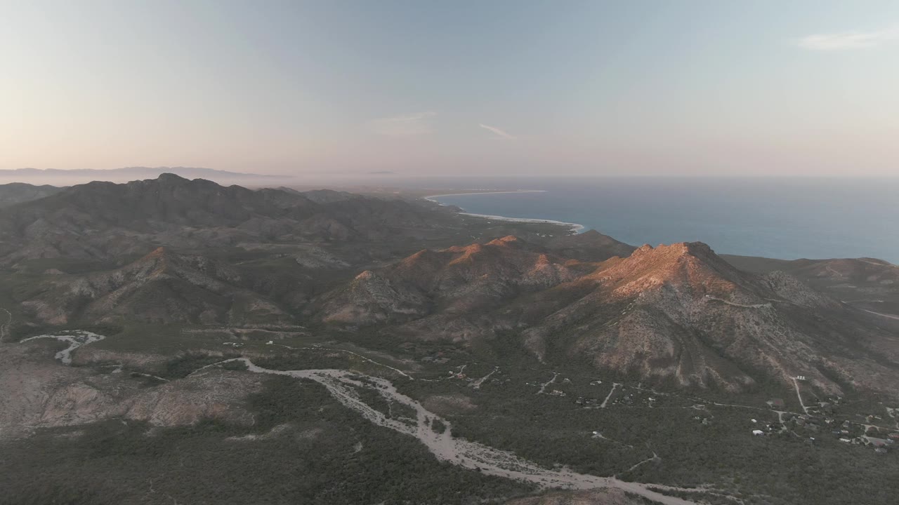 Cabo Pulmo, Baja California Sur, seen from above with layered dark ridges, a bright rocky peak at right, pale road traces across the valley, distant blue ocean, and no visible people.