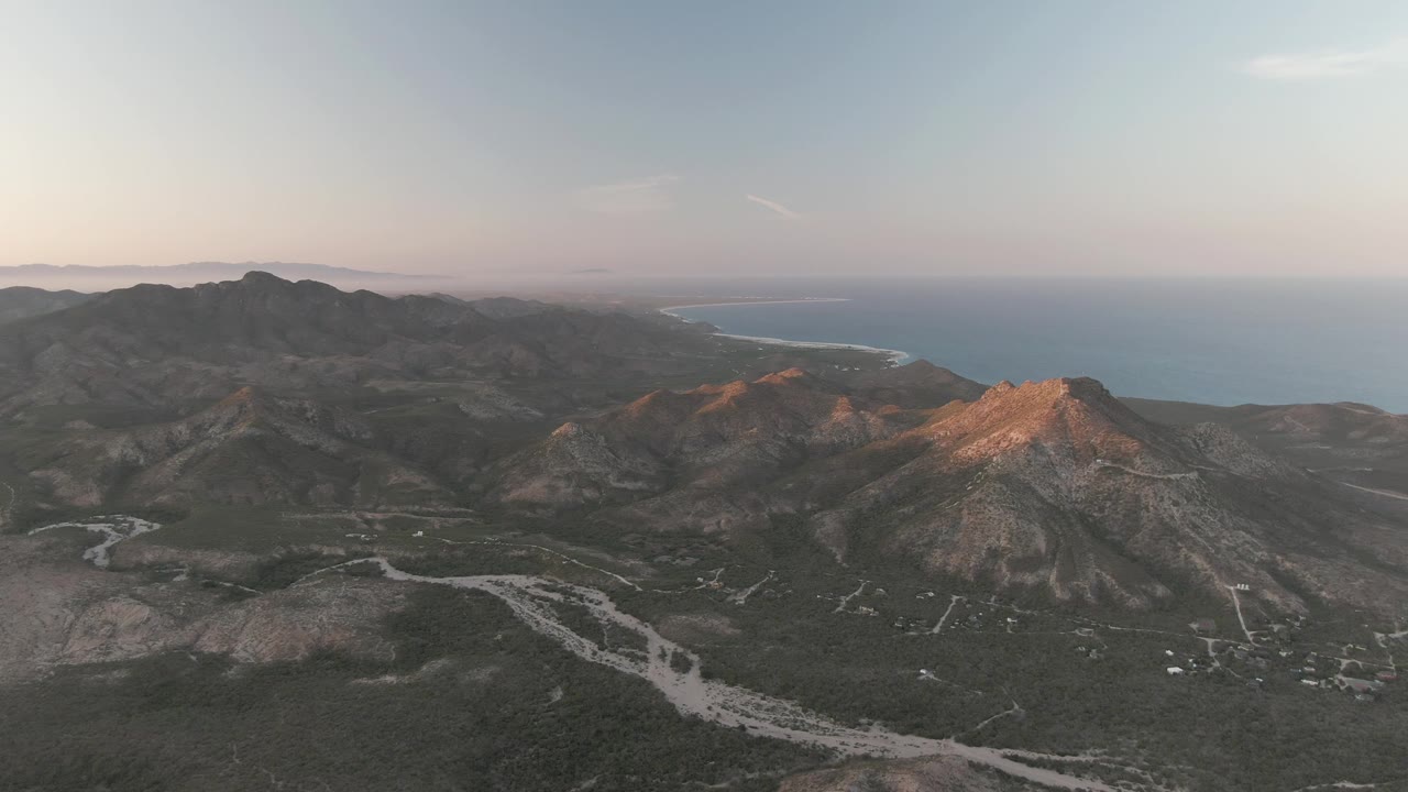 Mid-clip frame over Cabo Pulmo with the mountain mass centered left, a warmer lit ridge to the right, branching pale roads in the valley, distant ocean, and no visible people.