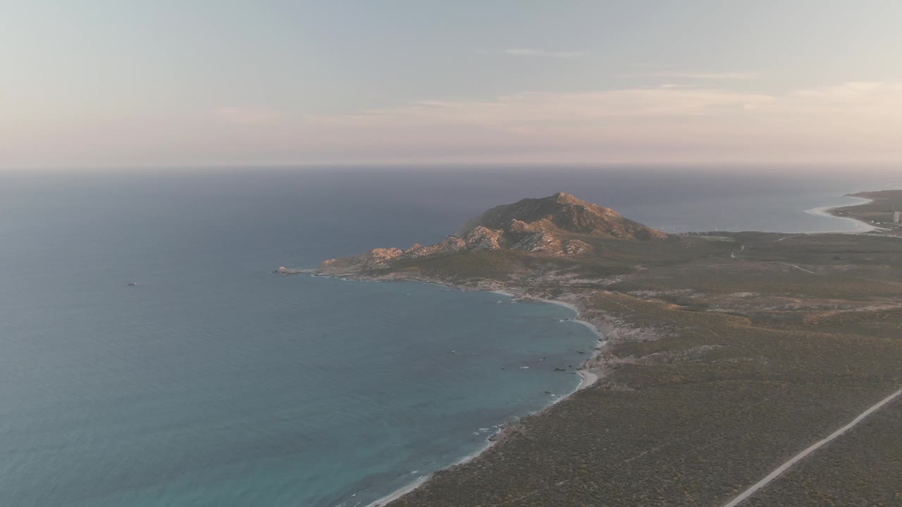 Cabo Pulmo aerial view at 0:01 with the curved shoreline below the hill, calm blue ocean filling the left side, a faint road at lower right, and no visible people.
