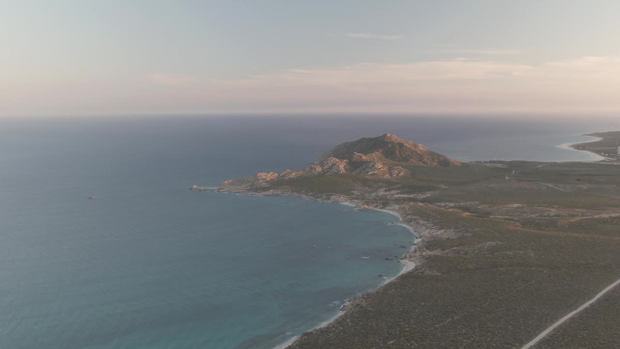 Mid-clip frame of Cabo Pulmo with the ocean occupying the left foreground, a pale road along the lower right scrubland, a rocky hill in the upper middle, and no visible people.