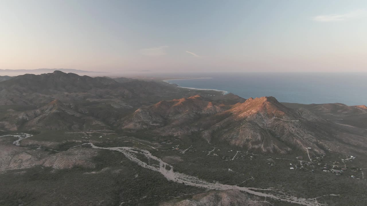 Aerial view of rugged mountains, winding pale road, and distant ocean horizon.