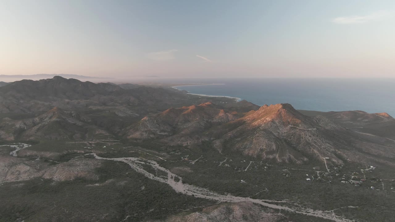 Cabo Pulmo aerial frame with dark mountain ridges across the background, pale tracks through the valley, a blue ocean strip beyond the land, and no visible people.