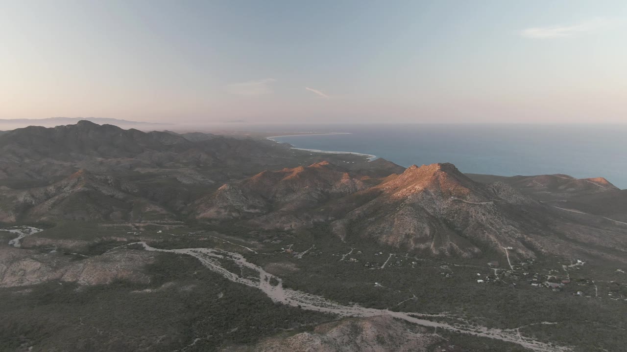 Aerial view in Cabo Pulmo with a rocky peak catching warm light, pale road lines across the valley floor, distant ocean and coastline, and no visible people.