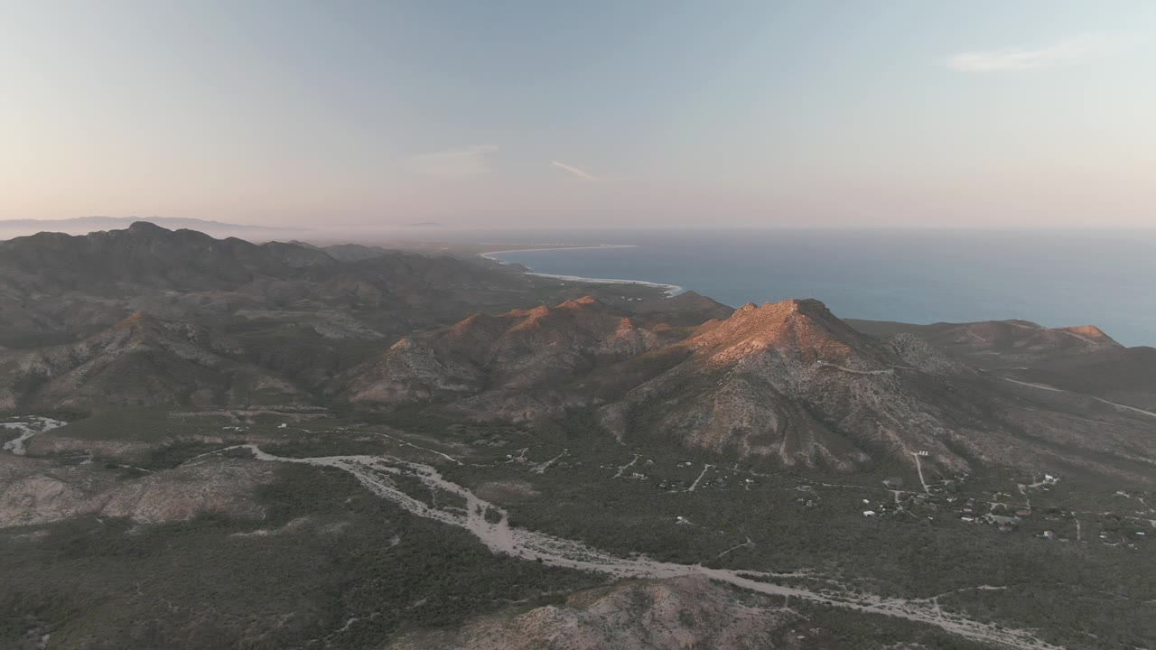 Wide Cabo Pulmo aerial frame showing a winding pale road or dry wash in the foreground valley, layered mountain slopes, blue ocean beyond the ridgeline, and no visible people.