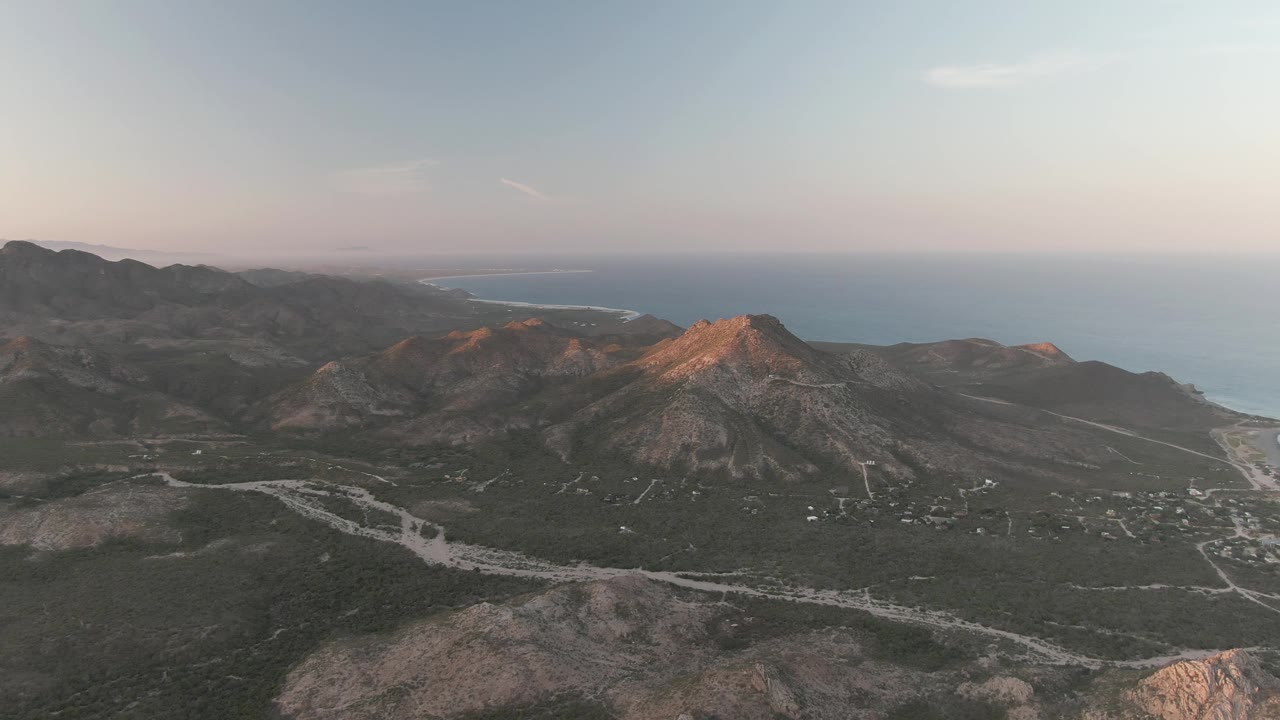 Opening aerial frame over Cabo Pulmo, Baja California Sur, with a rocky mountain ridge, pale winding roads through scrub terrain, distant ocean, and no visible people.