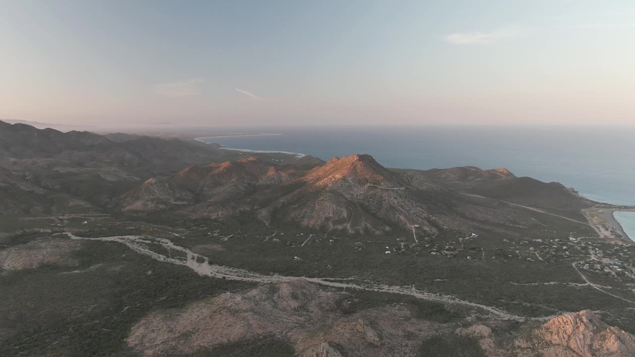 Wide drone-style frame over Cabo Pulmo showing rugged hills, a central sun-warmed peak, pale road lines through the valley, distant coastline, ocean, and no visible people.