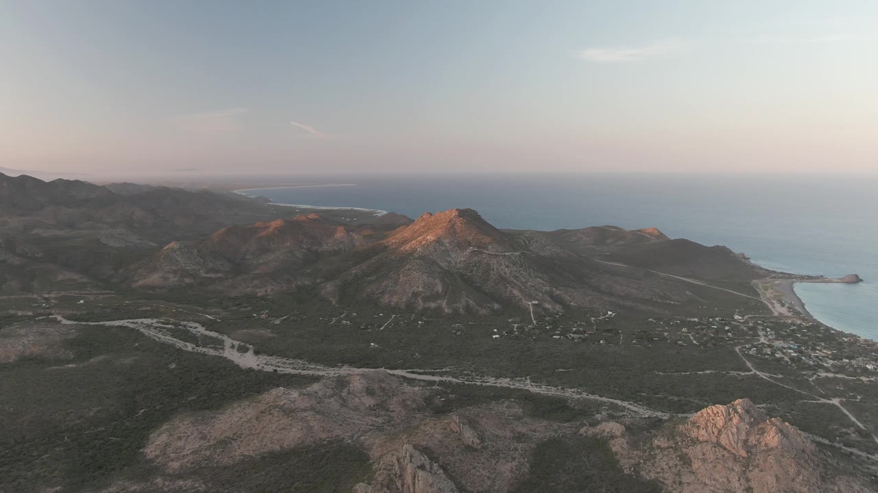 Aerial dusk view over Cabo Pulmo, Baja California Sur, with rocky ridges, winding roads, ocean on the horizon, and no visible people.