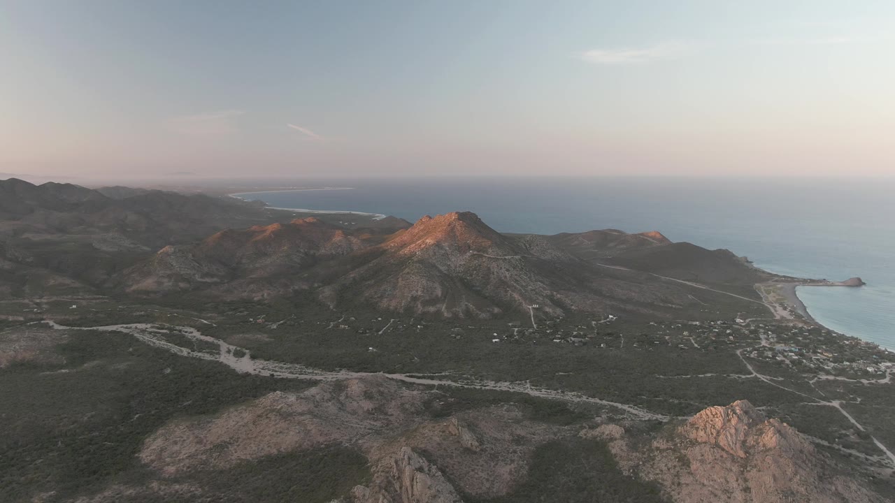 Wide aerial view in Cabo Pulmo showing a sunlit mountain ridge, darker valley terrain, shoreline roads, blue ocean, and no visible people.