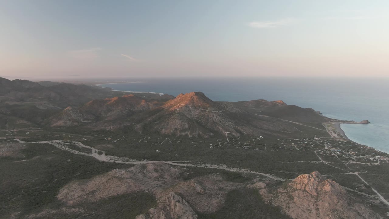Cabo Pulmo aerial frame with layered mountains, pale roads through dark vegetation, ocean and shoreline to the right, and no visible people.