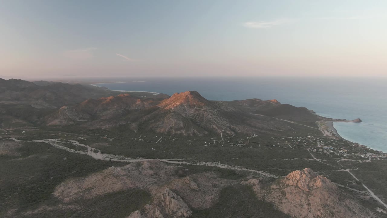 Dusk aerial of rocky hills, coastal valley, road lines, and ocean horizon.