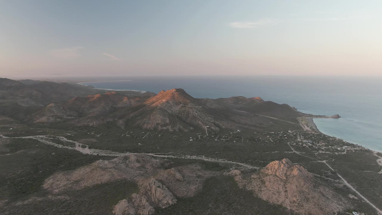 Aerial frame over Cabo Pulmo, Baja California Sur showing rocky hills, a pale road, blue ocean, soft sunset sky, and no visible people.