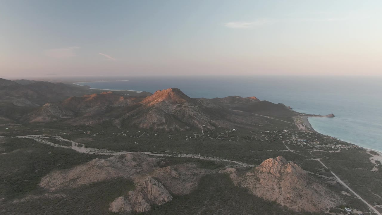 Aerial view in Cabo Pulmo with a winding road crossing dark green terrain, rocky peaks catching warm light, ocean at right, and no visible people.