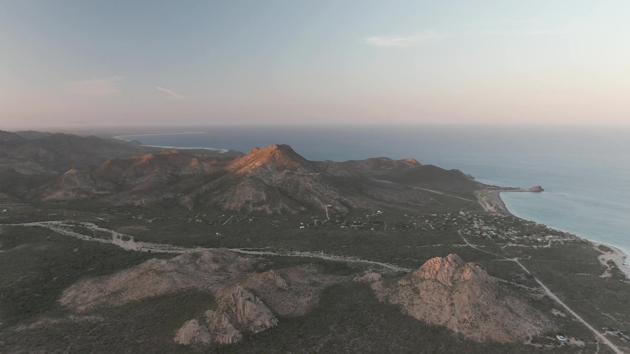 Drone-height view over Cabo Pulmo showing a sunlit peak, low rocky ridges, the coastal road network, ocean water, and no visible people.