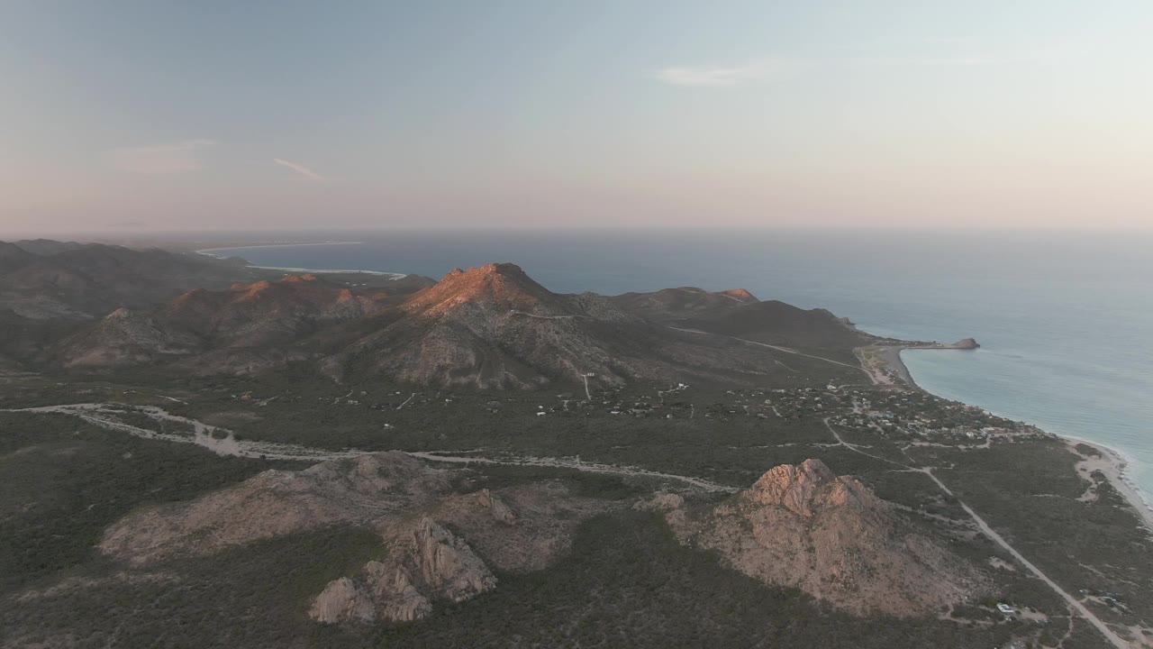Wide aerial of ocean shoreline, hills, and pale road.