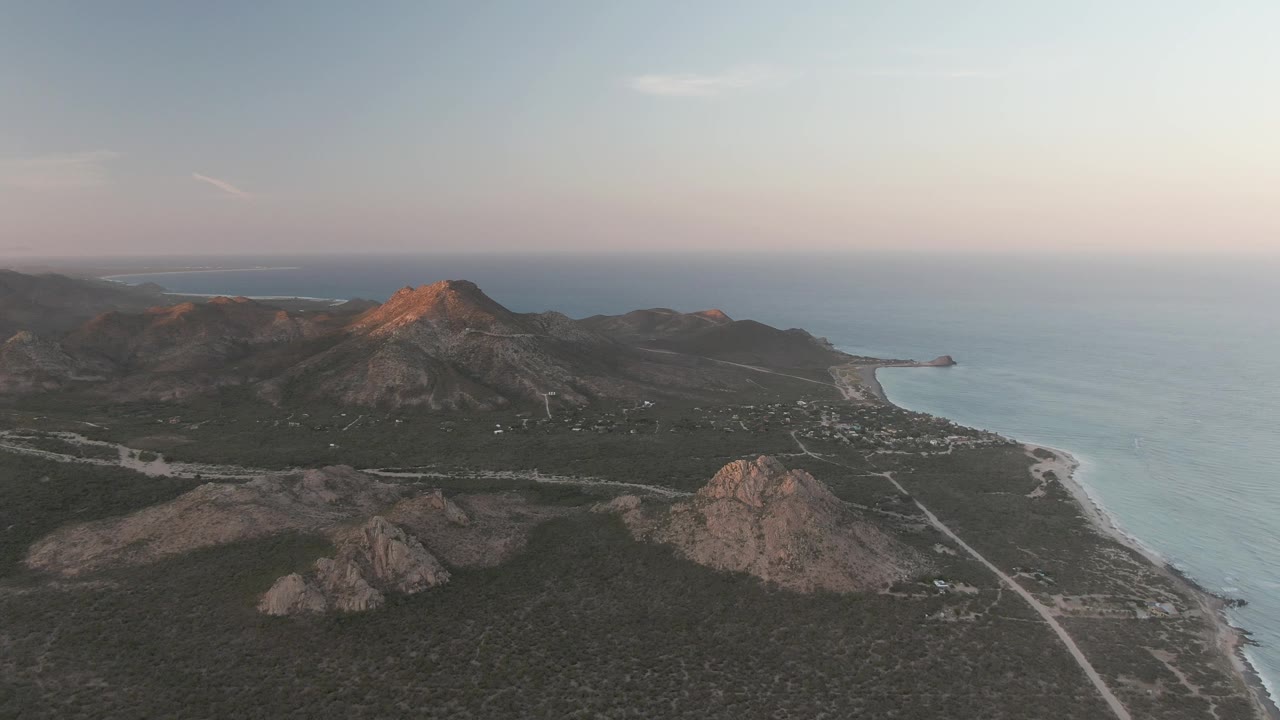 Aerial view over Cabo Pulmo rocky hills, sparse desert vegetation, a pale coastal road, and the ocean under soft sunset light, with no visible people.