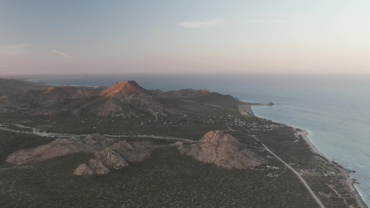 Wide Cabo Pulmo aerial showing the shoreline curving along the right side, a light road below the hills, dark scrub vegetation, and no visible people.