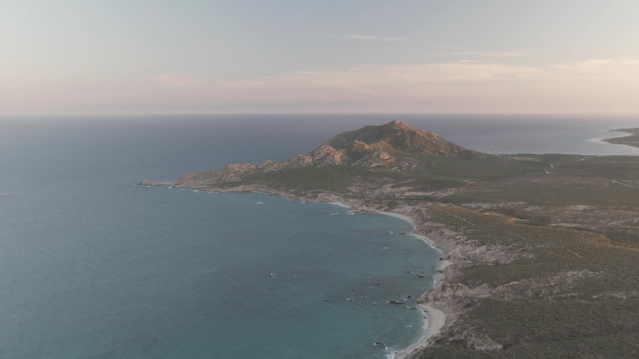 A wide Cabo Pulmo aerial frame shows the shoreline slightly farther along the curve, with small white surf marks, green-brown land, mountain ridge, and no visible people.