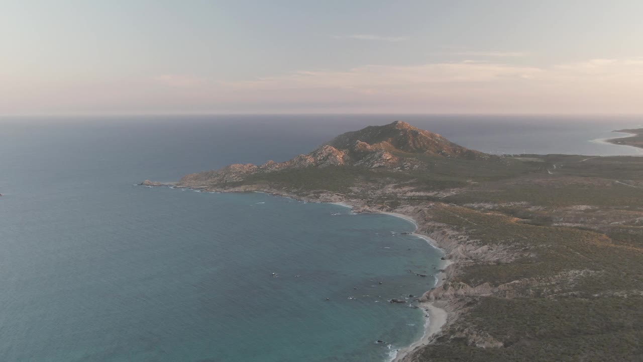 Cabo Pulmo coastline fills the frame with blue water at left, a narrow beach and surf line below, scrubland to the right, a rocky mountain beyond, and no visible people.