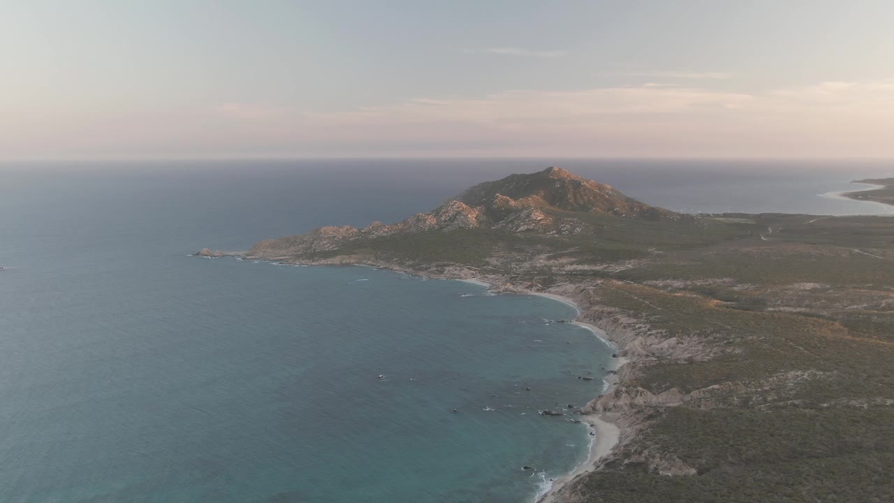 Later in the clip, the Cabo Pulmo aerial view shows soft light on the rocky mountain, darker coastal vegetation, shifting white foam along the beach, and no visible people.