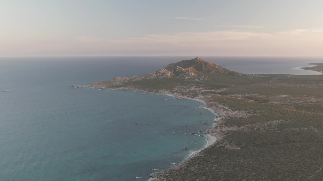 Aerial view over Cabo Pulmo with blue-green ocean on the left, a curved rocky and sandy shoreline, scrub-covered land, a distant coastal hill, and no visible people.