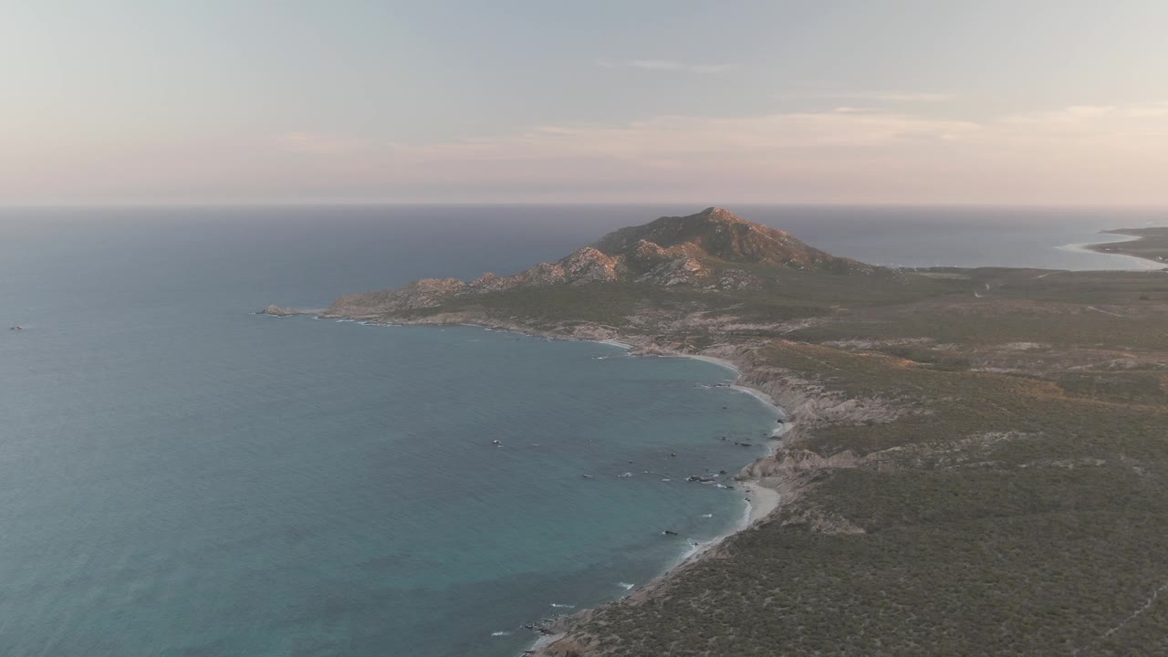 Wide aerial coastline with calm water, small boats, scrubland, and a hill.