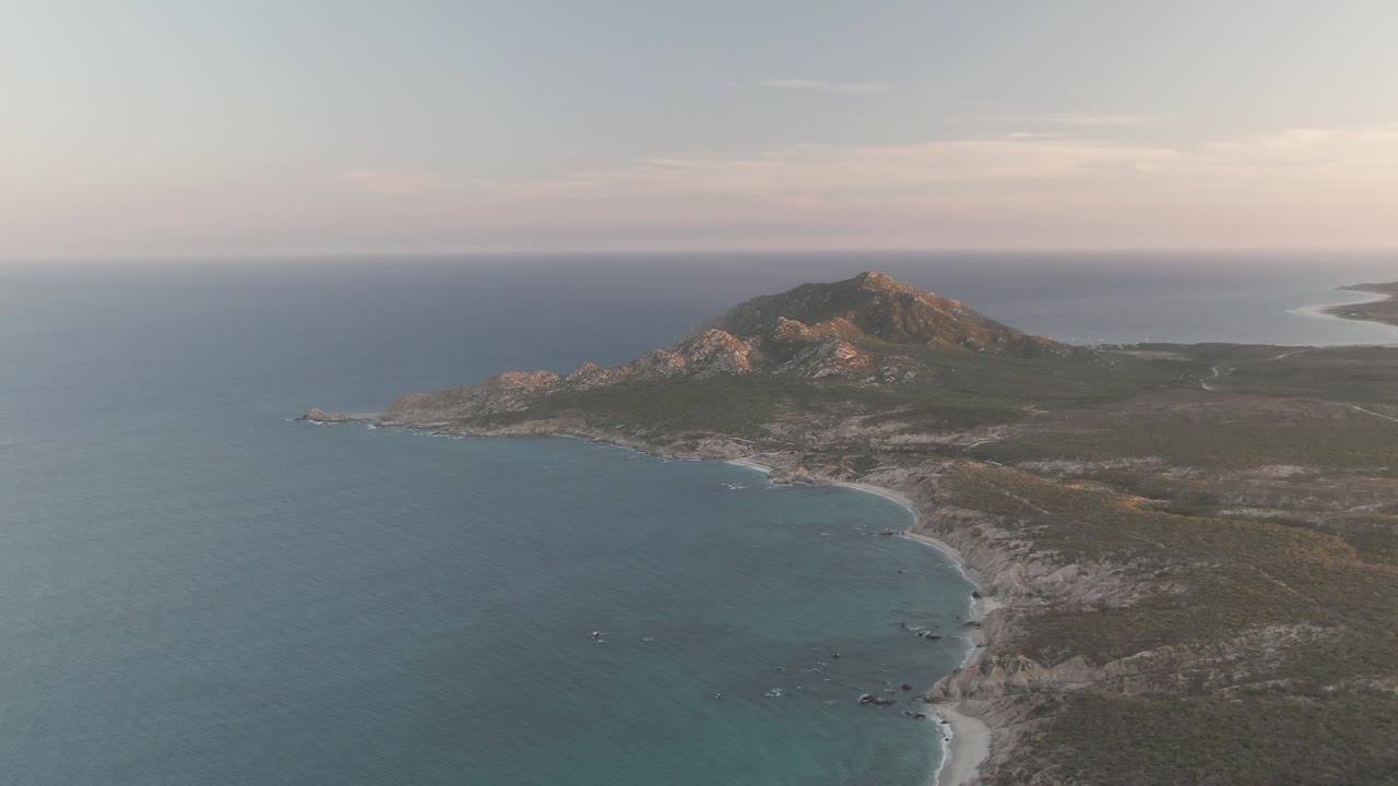Opening aerial frame over Cabo Pulmo with blue ocean in the foreground, a pale curving beach below the mountain, soft dusk clouds, and no visible people.
