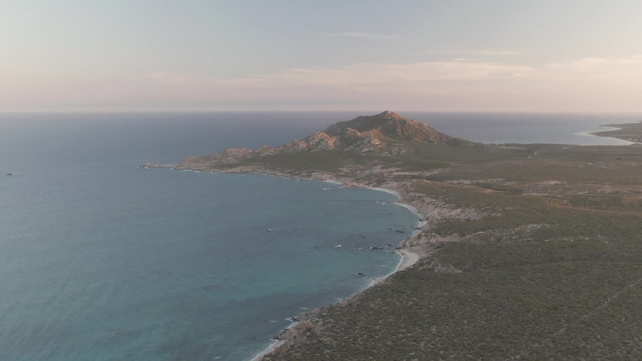 Opening frame over Cabo Pulmo, Baja California Sur, with turquoise water on the left, a pale shoreline below the hill, dry coastal terrain in the foreground, and no visible people.