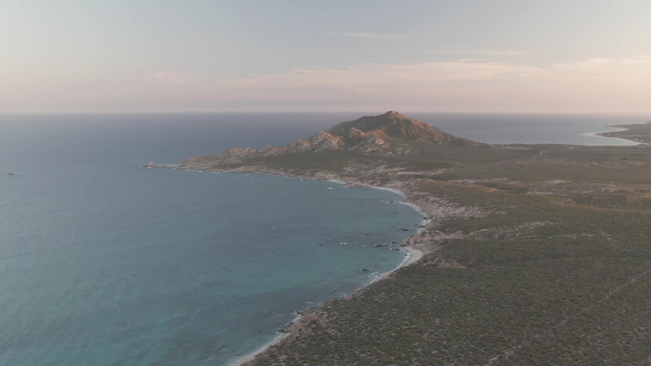 At 0:02, the aerial view shows the Cabo Pulmo beach curve more clearly beneath the rocky hill, with open sea to the left, dry land to the right, and no visible people.