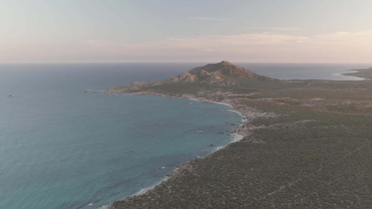 Aerial view of a curved beach, blue ocean, scrubland, and rocky hill.