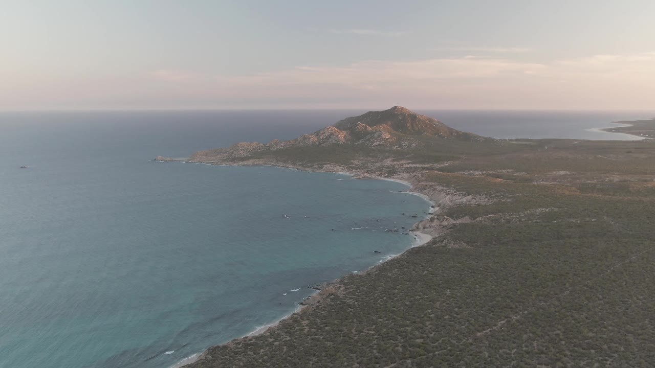 High aerial frame of Cabo Pulmo coastline, with the bay filling the left side, pale surf along the beach, dense scrubland to the right, the rocky hill centered in back, and no visible people.