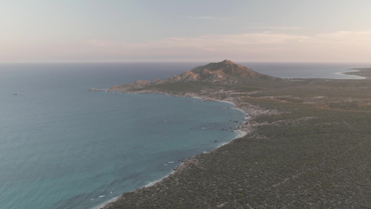 Cabo Pulmo aerial still with calm blue water stretching across the left foreground, a pale curved beach at midframe, scrub terrain on the right, a rocky hill beyond, and no visible people.