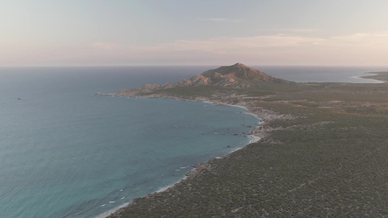 Aerial view of Cabo Pulmo coast with more open water on the left, small wave lines near the lower shore, scrubland covering the right side, a rocky hill near the horizon, and no visible people.