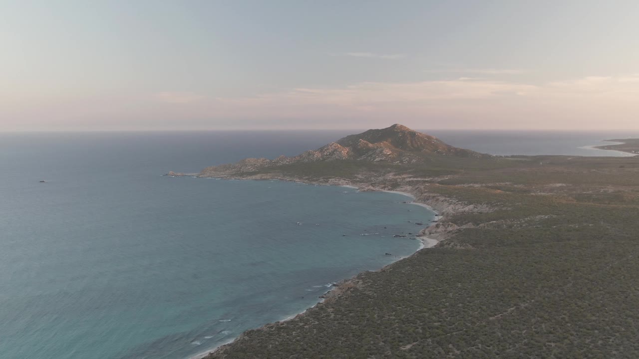 Aerial view of turquoise ocean, curving beach, coastal brush, and a rocky mountain.