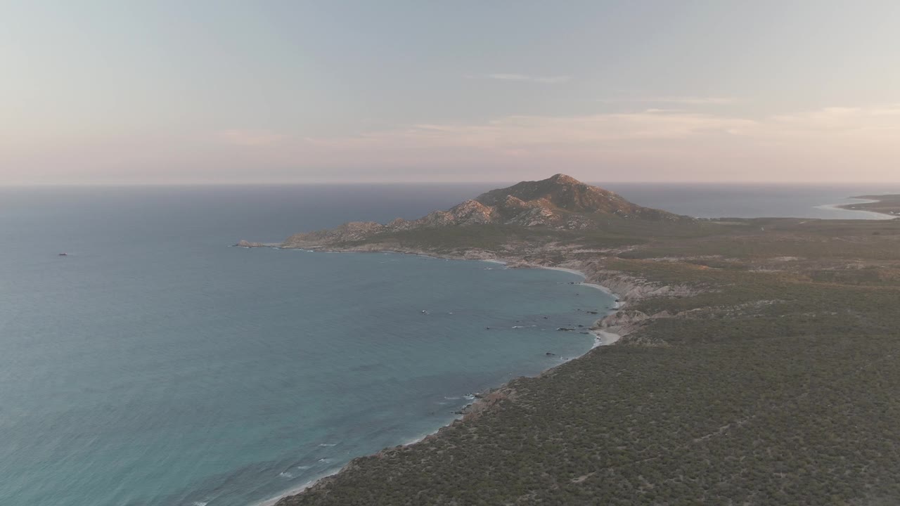 At 0:01, the Cabo Pulmo coastline remains in a high aerial composition with blue-green ocean, pale surf tracing the shore, low vegetation across the land, the mountain beyond, and no visible people.