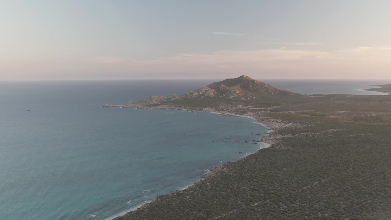 At 0:02, Cabo Pulmo appears from above with the open sea filling the left side, the beach curve cutting into the frame, sparse vegetation on the right, the mountain centered near the horizon, and no visible people.