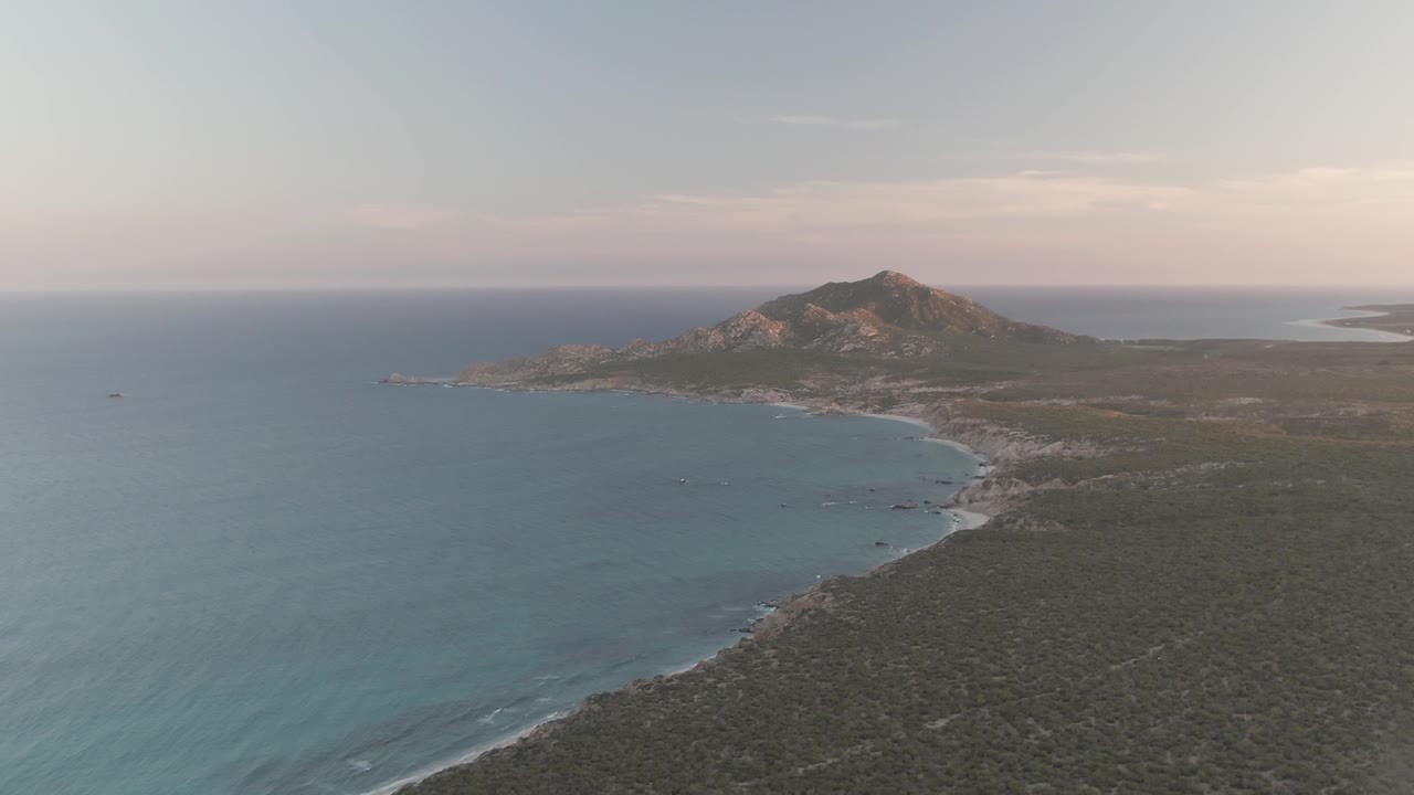 At 0:03, the aerial view of Cabo Pulmo shows calm turquoise water in the lower left, a narrow beach and rocky shore through the middle distance, dark coastal scrub in front, the mountain behind, and no visible people.