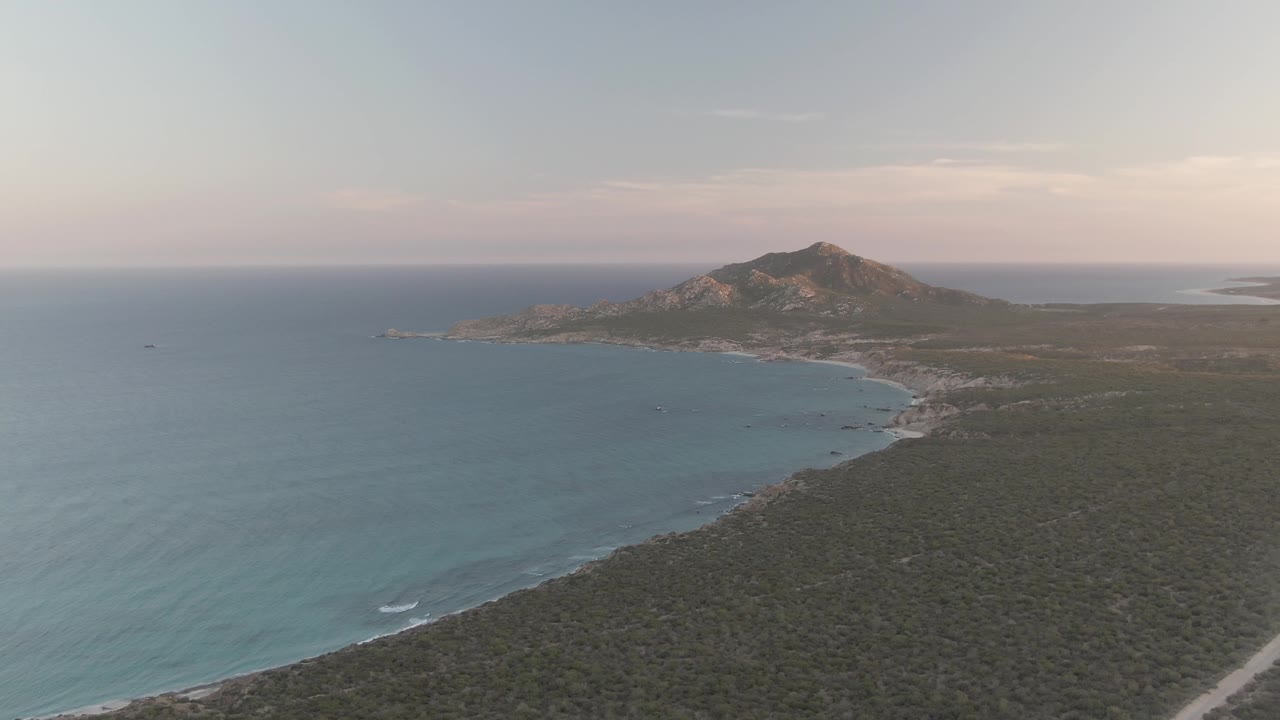 Wide aerial frame over Cabo Pulmo coastline with blue water on the left, dry scrubland below, a pale dirt road at lower right, a distant rocky hill, and no visible people.